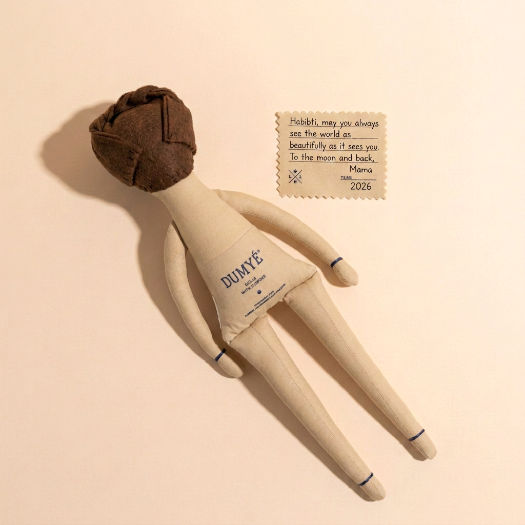 A handmade fabric doll with a brown fabric head and cream-colored body, lying on a beige background next to a small printed card addressed to 'Mama'.