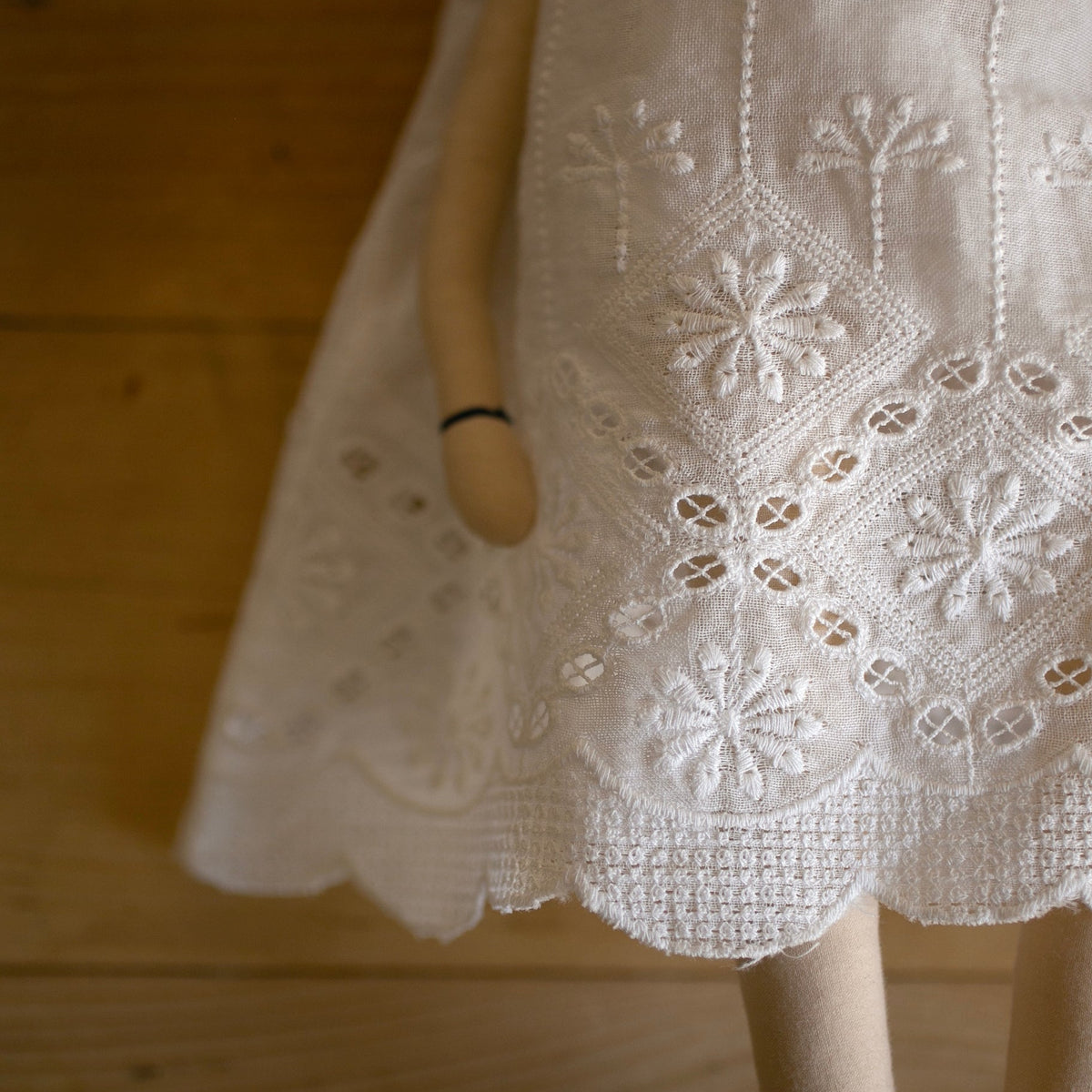 A close-up of a white embroidered dress or skirt with floral eyelet embroidery, shown on a heirloom doll against a warm wood-paneled background.